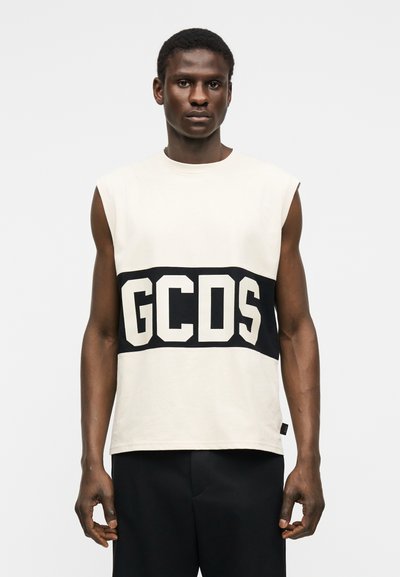 Άνδρας που φοράει λευκό αμάνικο τοπ με φαρδιά μαύρη λωρίδα και μεγάλα λευκά γράμματα "GCDS", συνδυασμένο με μαύρο παντελόνι, στέκεται μπροστά σε μονόχρωμο φόντο.