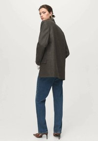 Hnědý tvídový blazer s uvolněným střihem, v kombinaci s rovnými modrými džíny a hnědými lodičkami na vysokém podpatku; modelka zády k fotoaparátu.