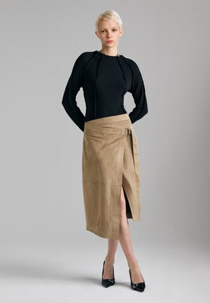 Donna con capelli biondi corti che indossa una maglia nera aderente a maniche lunghe, gonna beige a portafoglio con spacco laterale e tacchi neri appuntiti, in piedi davanti a uno sfondo grigio.