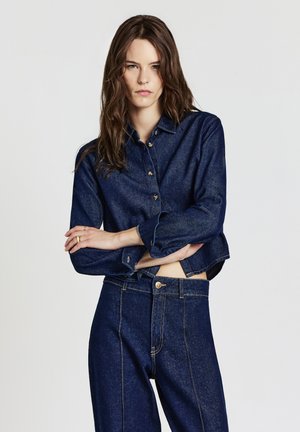 Femme aux longs cheveux bruns portant une chemise en denim bleu foncé et un pantalon en denim taille haute assorti, se tenant les bras croisés contre un fond uni.