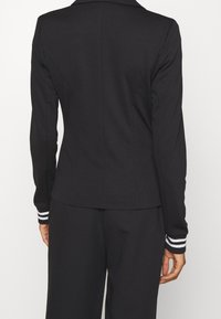 Blazer noir avec des manches longues, fabriqué en tissu lisse, présentant un design ajusté et des accents rayés blancs sur les poignets.