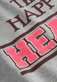 Grauer Stoff mit dem strukturierten, rosa bestickten Schriftzug "HAPPY" und dunkelbraunem Text darüber. Zeigt horizontale braune Streifen auf dem grauen Hintergrund.