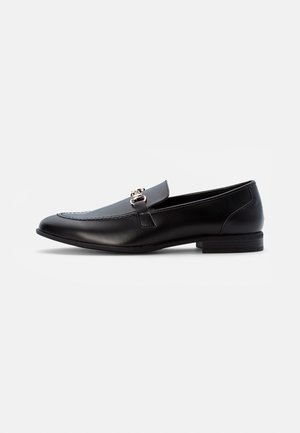 Mocassins en cuir noir avec bout arrondi, détails cousus et un accent de ferrure dorée à l'avant. Talon bas empilé.