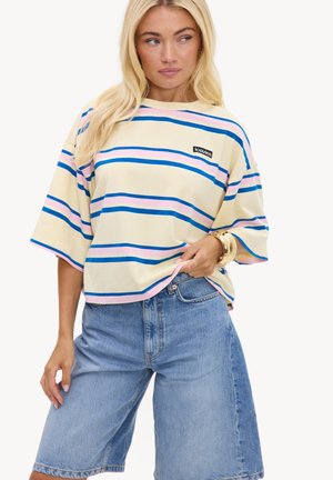 Femme portant un t-shirt crème oversize avec des rayures bleues et roses et un short long en denim bleu clair, tenant l'ourlet du t-shirt d'une main.