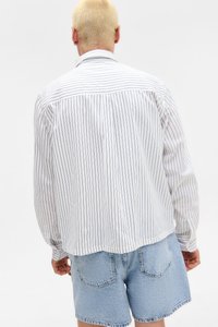 Person med kort blondt hår iført en hvit langermet skjorte med tynne svarte striper og lyseblå denimshorts, vendt bort.