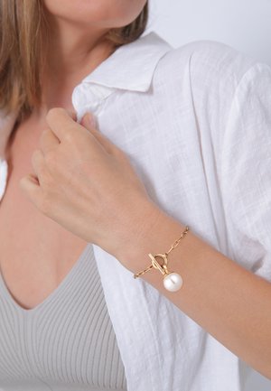 Goldene Kettenarmband mit einer Perle und einem Toggle-Verschluss, getragen an einem Handgelenk mit einem hellen Oberteil und einem weißen Leinenhemd.