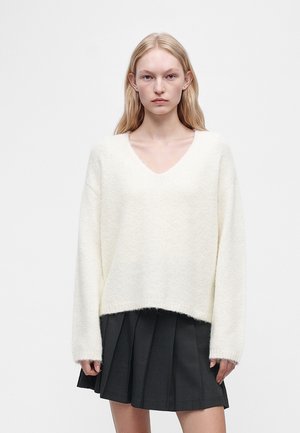 Jeune femme aux longs cheveux blonds portant un pull blanc doux à col en V et une jupe plissée noire sur un fond uni.