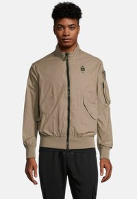 Beige bomberjack met een rits aan de voorkant, ribgebreide manchetten en kraag. Beschikt over twee zijzakken en een ritszak op de linkermouw.