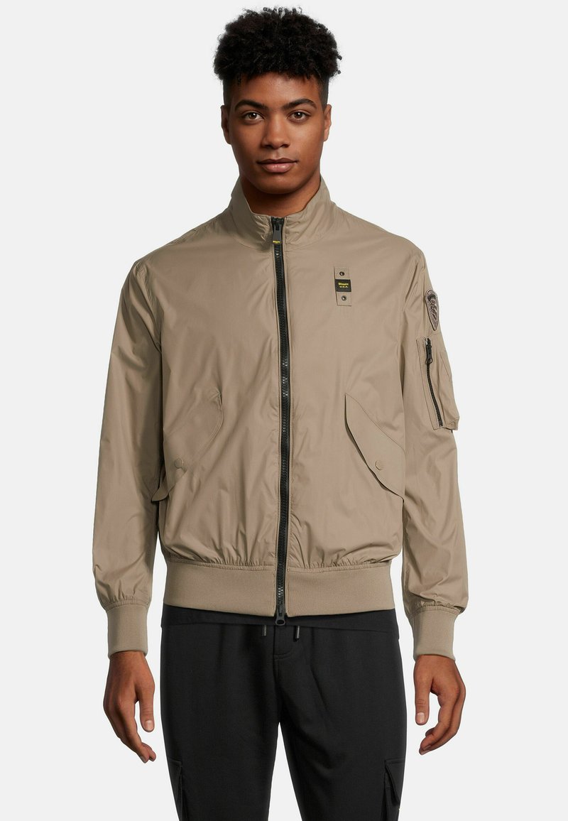 Beige bomberjack met een rits aan de voorkant, ribgebreide manchetten en kraag. Beschikt over twee zijzakken en een ritszak op de linkermouw.