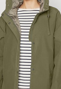 Oliwkowa kurtka z kapturem, z zapięciem na zamek, dwoma bocznymi kieszeniami i napami, nałożona na białą koszulę w niebiesko-białe paski.