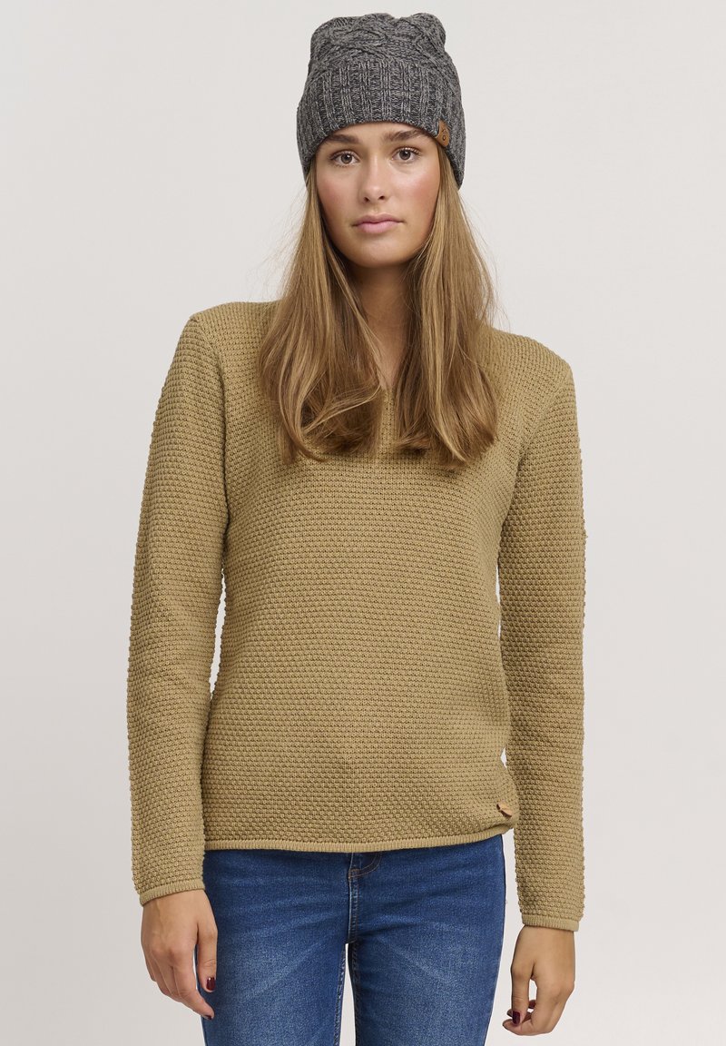 Maglione beige a trama con maniche lunghe, scollatura rotonda, design aderente e piccolo dettaglio in pelle. Indossato con jeans blu.