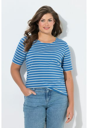 Vrouw met lang golvend haar, die een blauw-wit gestreept T-shirt met korte mouwen en lichtblauwe spijkerbroek draagt, staat met één hand in de zak.