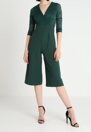 Femme portant une combinaison vert foncé à manches en dentelle avec un pantalon large raccourci et des sandales noires à talons hauts avec bride à la cheville, debout sur un sol blanc.