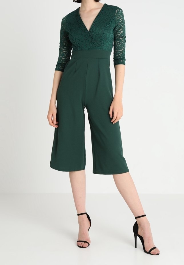 Femme portant une combinaison vert foncé à manches en dentelle avec un pantalon large raccourci et des sandales noires à talons hauts avec bride à la cheville, debout sur un sol blanc.