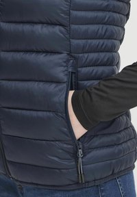 Mornarska pufasta prsluk s horizontalnim šavovima, prednjim džepovima na zip, i glatkom završnom obradom. Ispod se nosi crna majica dugih rukava.