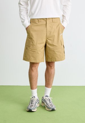 Beige cargo shorts with two side pockets, a back pocket, and a small logo on the front. Worn with grey patterned sneakers and white socks.