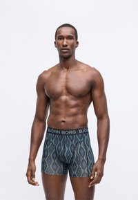 Muskuløs mann står, iført mønstrede blågrønne Björn Borg boxershorts, mot en ensfarget hvit bakgrunn.