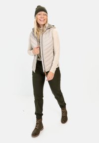 Helle graue gesteppte Weste mit Kapuze, beige Langarmshirt, olivgrüne Hose und braune Schnürstiefel. Model lächelt vor einem einfarbigen Hintergrund.