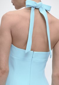 Πλάτη ενός ατόμου που φοράει ανοιχτό μπλε φόρεμα με λουράκι στον λαιμό δεμένο με φιόγκο και ορατό φερμουάρ στο κέντρο της πλάτης.