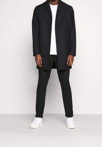 Manteau long marine avec revers crantés, trois boutons, porté sur un t-shirt blanc et un pantalon noir, complété par des baskets blanches.