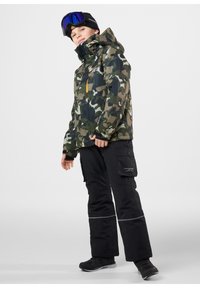 Kamouflagejacka i nyanser av grönt med dragkedjefickor, ihop med svarta byxor med cargofickor och reflexränder. Svart fotbeklädnad ingår.