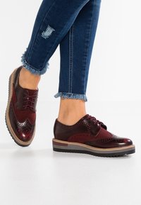 Chaussures en cuir et suède bordeaux à lacets avec des détails perforés, une semelle texturée et des accents marron contrastants. Portées avec des jeans à ourlet effiloché.