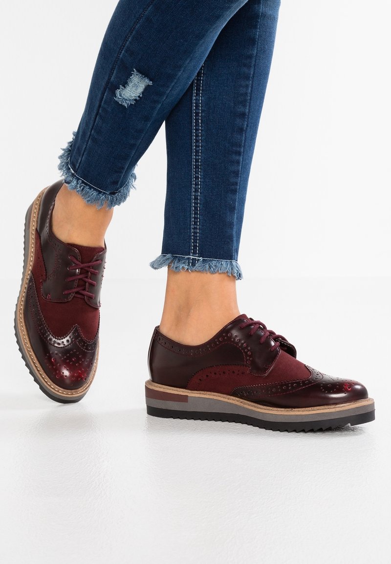 Chaussures en cuir et suède bordeaux à lacets avec des détails perforés, une semelle texturée et des accents marron contrastants. Portées avec des jeans à ourlet effiloché.