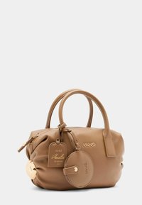 Sac à main beige avec deux poignées, une fermeture éclair et des accents dorés. Comprend une étiquette de marque et un charme rond attaché. Surface lisse et texturée.