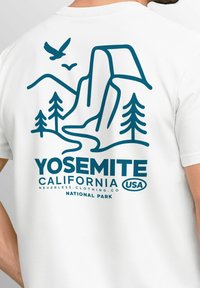 Weißes T-Shirt mit einem blauen grafischen Design der Berge, Bäume und Vögel von Yosemite, mit dem Text "YOSEMITE CALIFORNIA USA NATIONAL PARK."