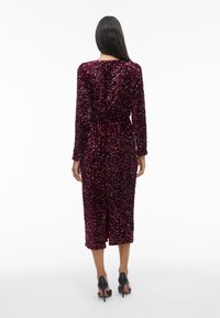 Kvinne iført en burgunder midi-kjole med lange ermer og paljetter, med splitt i ryggen, kombinert med svarte høyhælede sko, står mot en hvit bakgrunn.