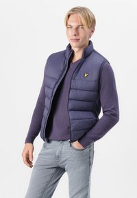 Gilet en duvet bleu marine avec un design matelassé, fermeture éclair à l'avant et col montant, porté par-dessus un pull violet, associé à un jean gris clair. Détail logo jaune.