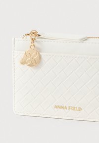 Porte-cartes en texture tissée blanche avec des accents dorés, une fermeture éclair et un charm décoratif à motif floral. Marque : ANNA FIELD.