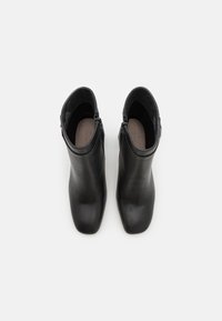 Un par de botas de cuero negro hasta el tobillo con puntas cuadradas y cremalleras en los lados internos, vistas desde arriba sobre un fondo blanco.