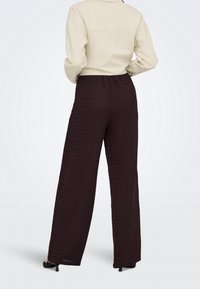 Personne portant un pantalon marron foncé texturé, taille haute, à jambes larges, avec des talons noirs et une chemise beige à manches longues rentrée, vue de dos.
