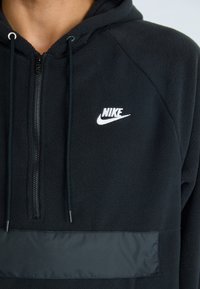 Črna flis majica s kapuco z zadrgo spredaj, vsebuje izrazit bel logotip Nike in kontrastno gladko blago ploščo čez prsi.