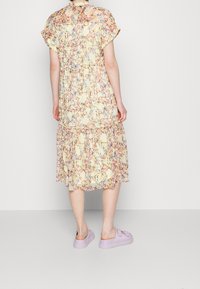 Personne portant une robe midi jaune à fleurs avec des manches courtes et des baskets lilas à enfiler, se tenant devant un fond blanc uni.