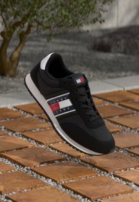 Zwarte atletische sneaker met een gladde textuur, voorzien van blauwe, witte en rode strepen met de naam "TOMMY JEANS" aan de zijkant, en een witte zool.