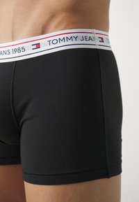 Boxer ajustados negros con una cinturilla blanca que presenta acentos en rojo y azul marino. La cinturilla está marcada con "TOMMY JEANS 1985" en una fuente en negrita.