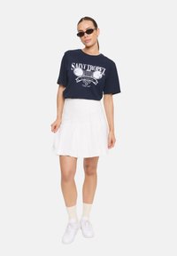 T-shirt graphique marine avec impression "Saint Tropez", associé à une jupe plissée blanche, des sneakers blanches et des lunettes de soleil noires.