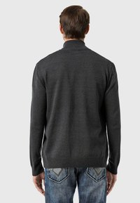 Dunkelgrauer gestrickter Pullover mit Stehkragen, langen Ärmeln und geripptem Saum, kombiniert mit blauen Jeans, die über auffällige Verzierungen an den Gesäßtaschen verfügen.