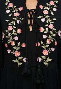 Μαύρο ένδυμα με ροζ και κόκκινη κεντητική με λουλούδια, κορδόνια δέσιμου στο ντεκολτέ και μαύρα φουντωτά κορδόνια μπροστά.