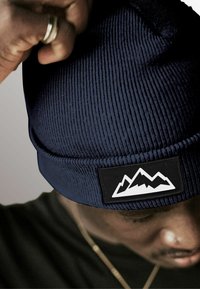 Navy blue knitted beanie with ribbed texture and a black patch featuring a white mountain design. The cuff is foldable.