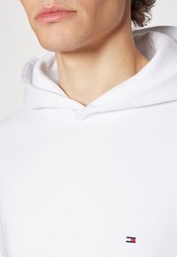 Hoodie blanc avec une texture légère et douce. Il présente une capuche avec cordon de serrage et un petit logo brodé en rouge, blanc et bleu au bas.