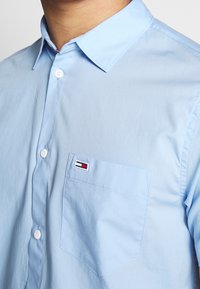 Ljusa blå, knapp-up skjorta gjord av mjuk bomull. Har en vänster bröstficka med en liten logotyp i röd, vit och blå sömnad.