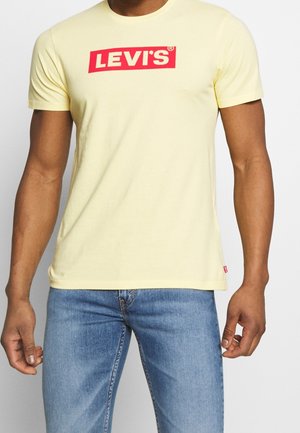 Mężczyzna w żółtej koszulce Levi's z czerwonym logo i niebieskich dżinsach, stojący na jednolitym białym tle.