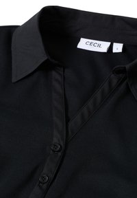 Chemise noir à boutons avec un col en V, fabriquée en tissu lisse. Contient des boutons noirs et une étiquette indiquant "CECIL" taille "S."
