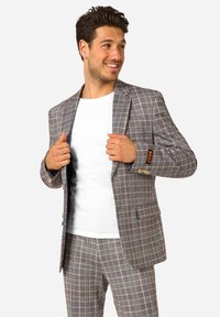 Blazer de cuadros gris con botones dorados, que presenta dos bolsillos delanteros y un puño de marca en contraste. Llevado sobre una camiseta blanca.