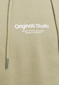 Khaki hoodie met witte tekst "Originals Studio" en zilveren trekkoordaccenten; gemaakt van zachte, gestructureerde stof.