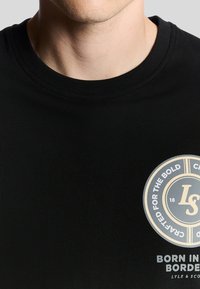 T-shirt en coton noir avec un col rond, arborant un logo circulaire en texte doré et blanc sur le devant. Texture lisse, design ajusté.