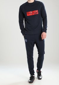 Felpa blu navy con "PARIGI" in rosso sul petto, abbinata a pantaloni jogging coordinati con un logo e scarpe da ginnastica nere.